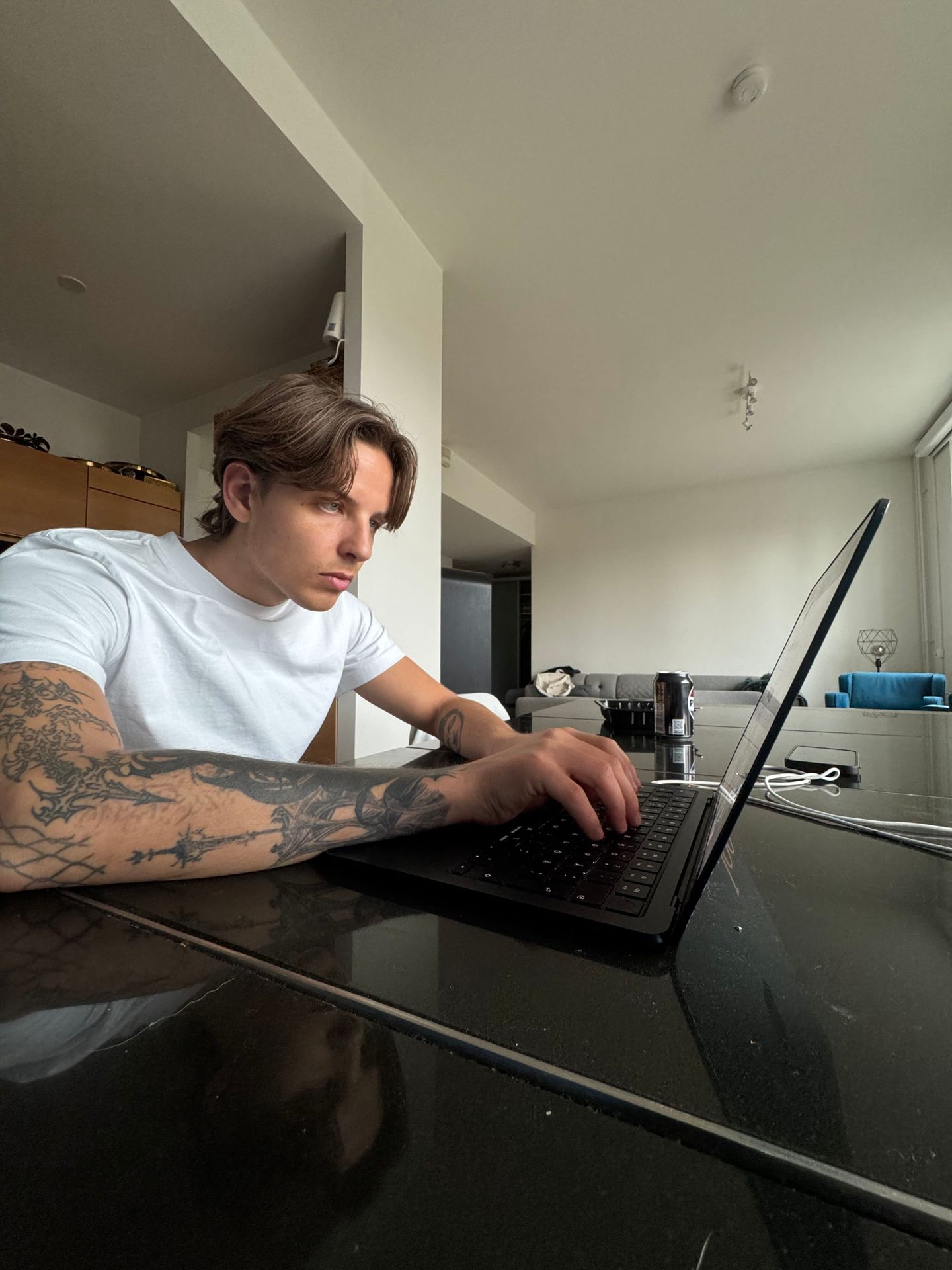 A young man with tattooed arms sits at a table and types on a laptop in a bright, modern living space.