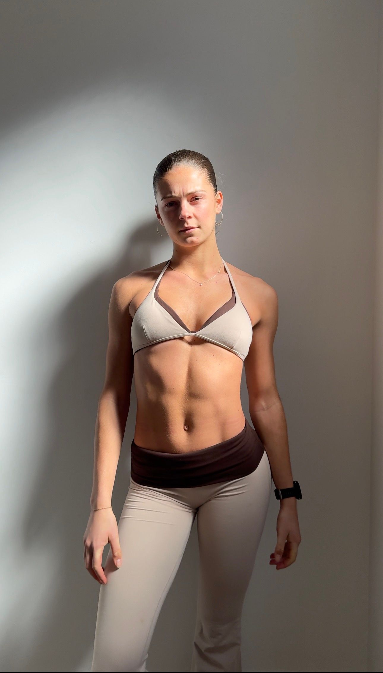 A fit woman with her hair pulled back stands against a plain wall wearing a beige sports bra and matching leggings.