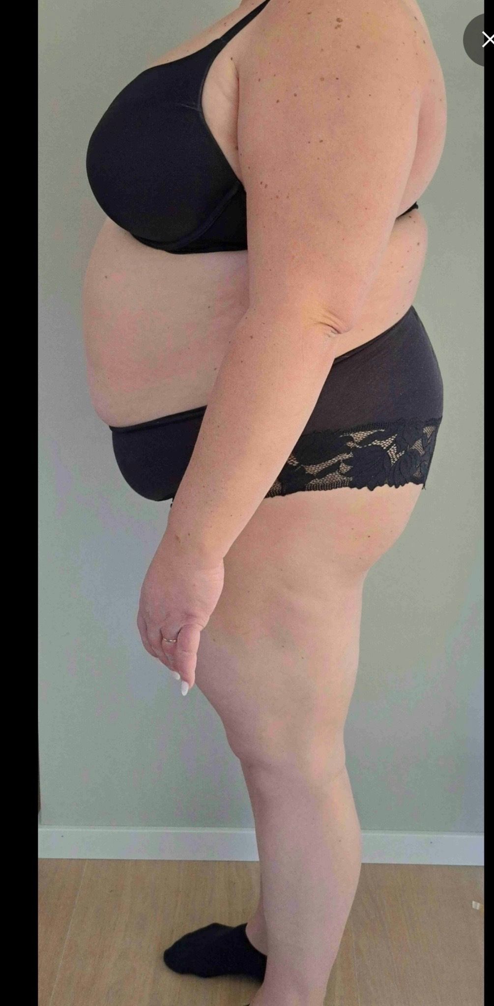 A person stands in profile wearing black undergarments against a plain wall.