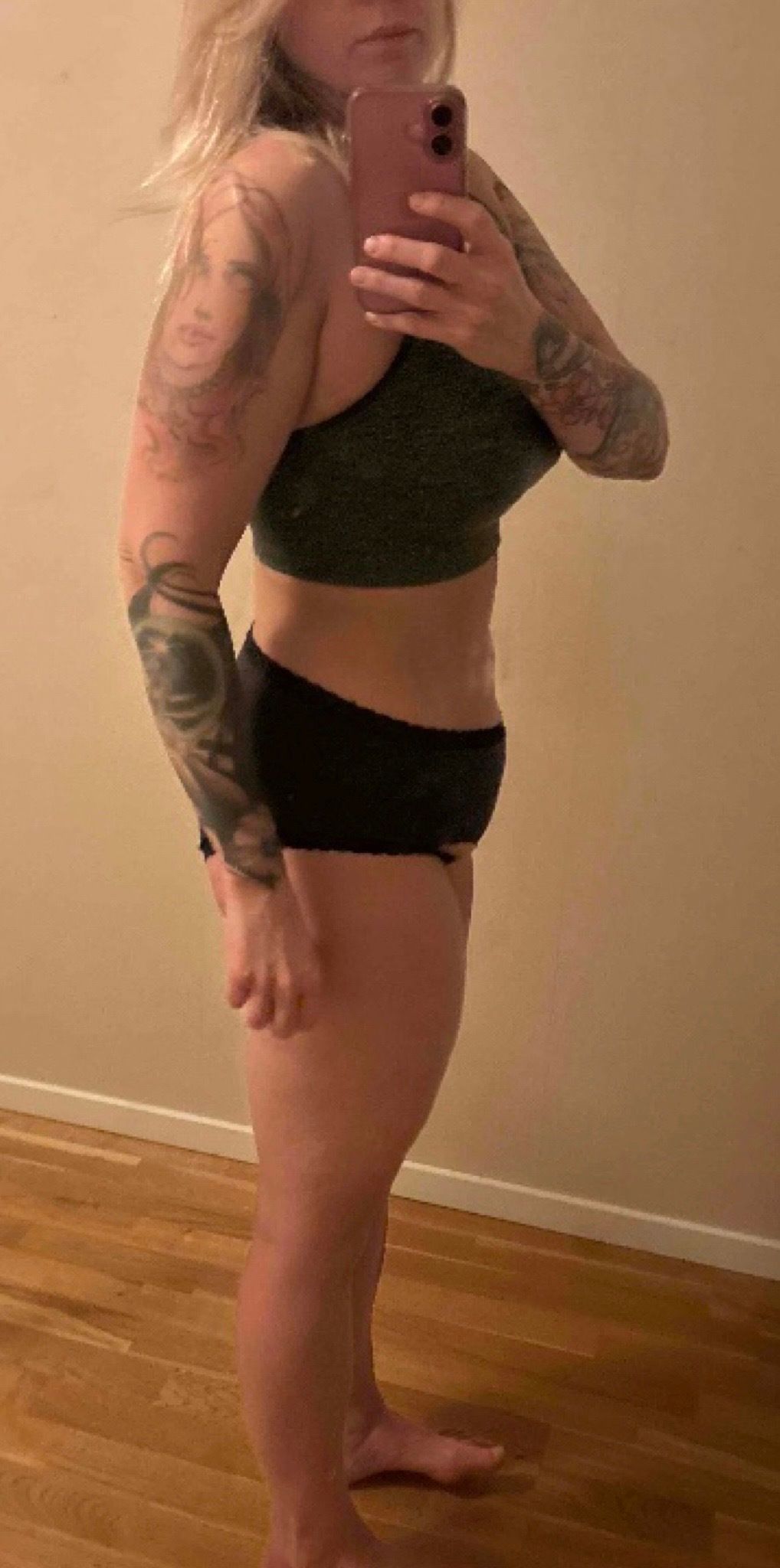 A woman with arm tattoos takes a side-profile mirror selfie in a sports bra and underwear.