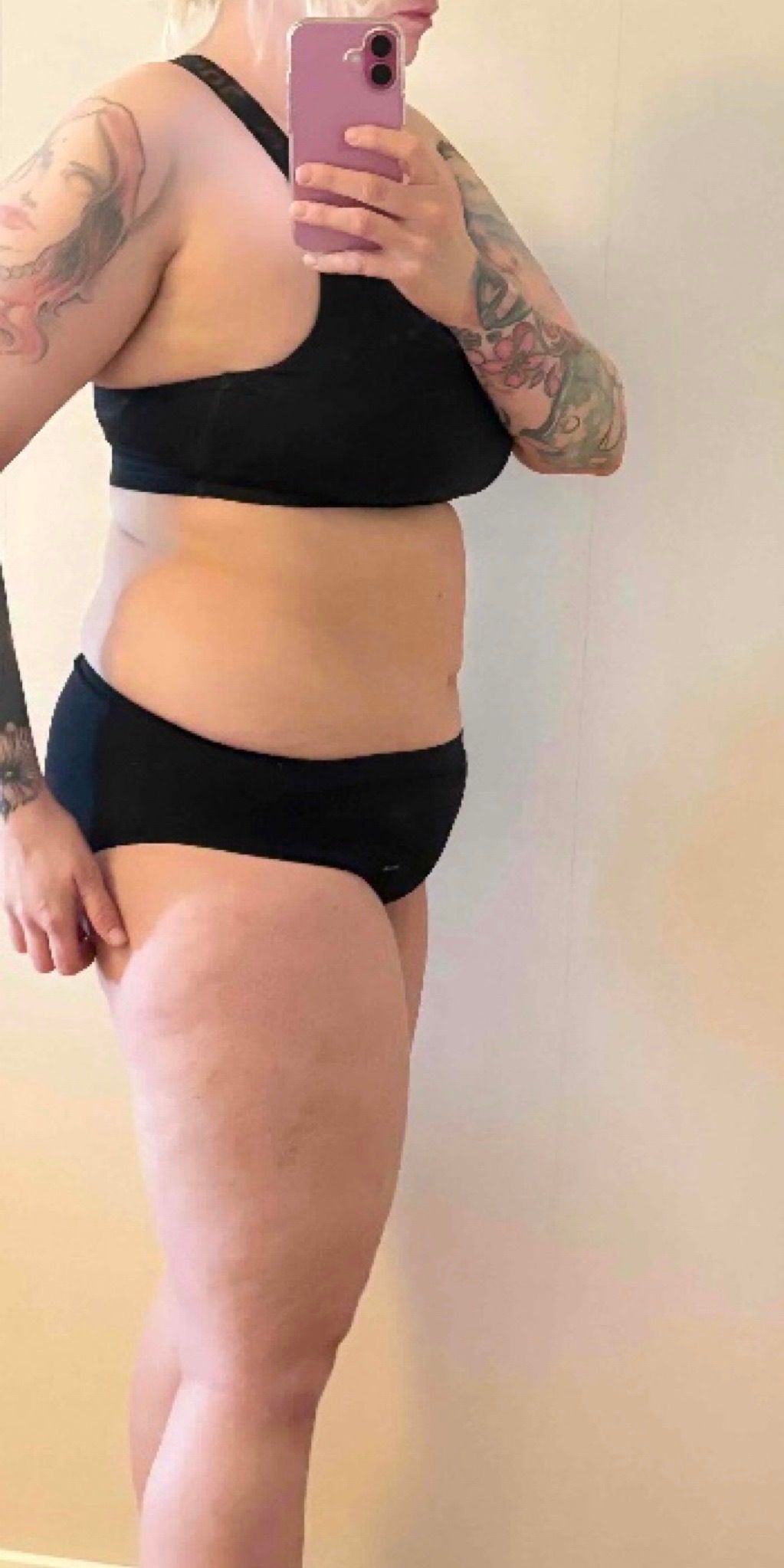 A person with tattoos stands in profile wearing black athletic wear while taking a mirror selfie.
