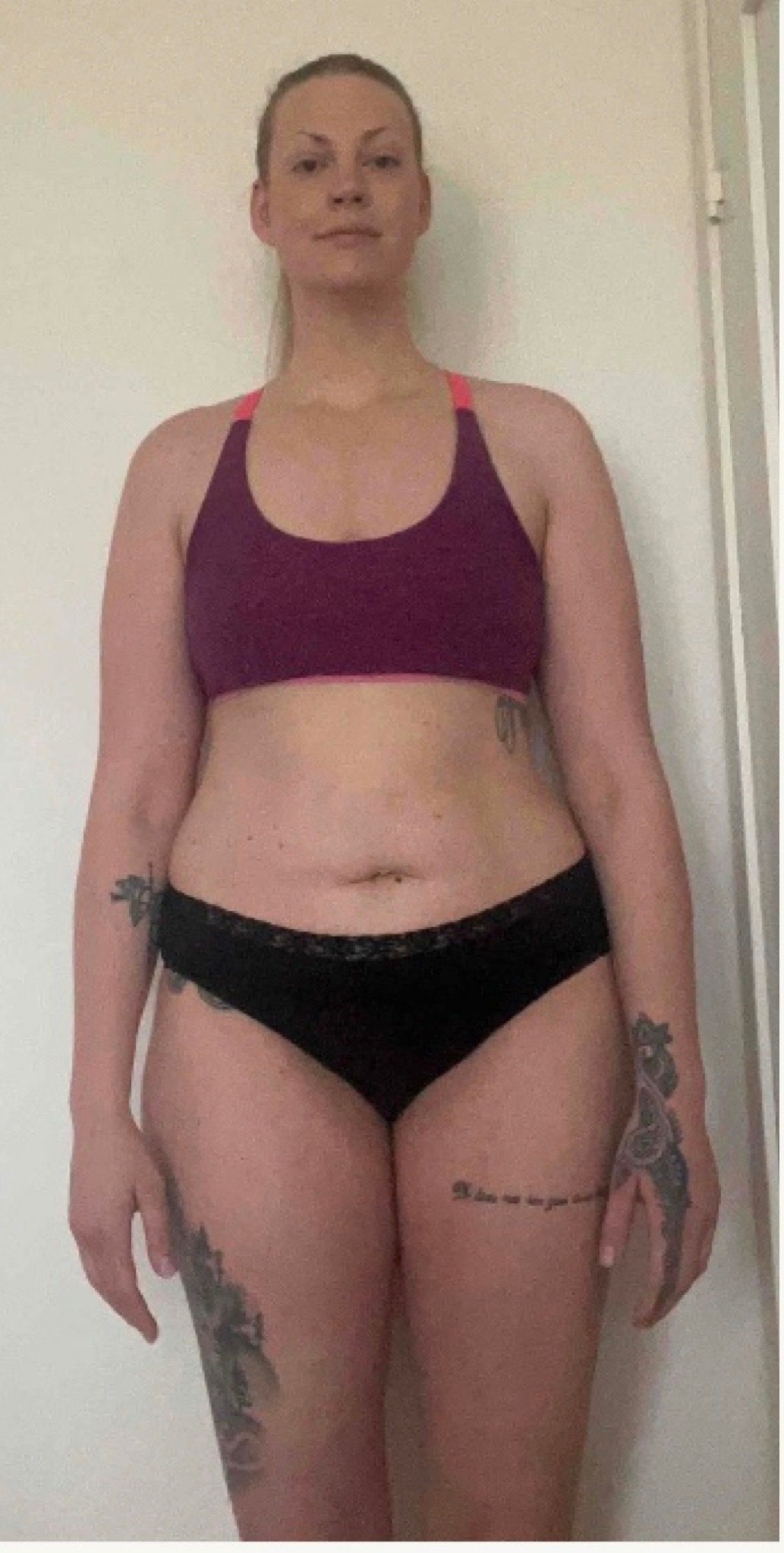 A woman with tattoos stands facing forward against a plain wall, wearing a purple sports bra and black underwear.