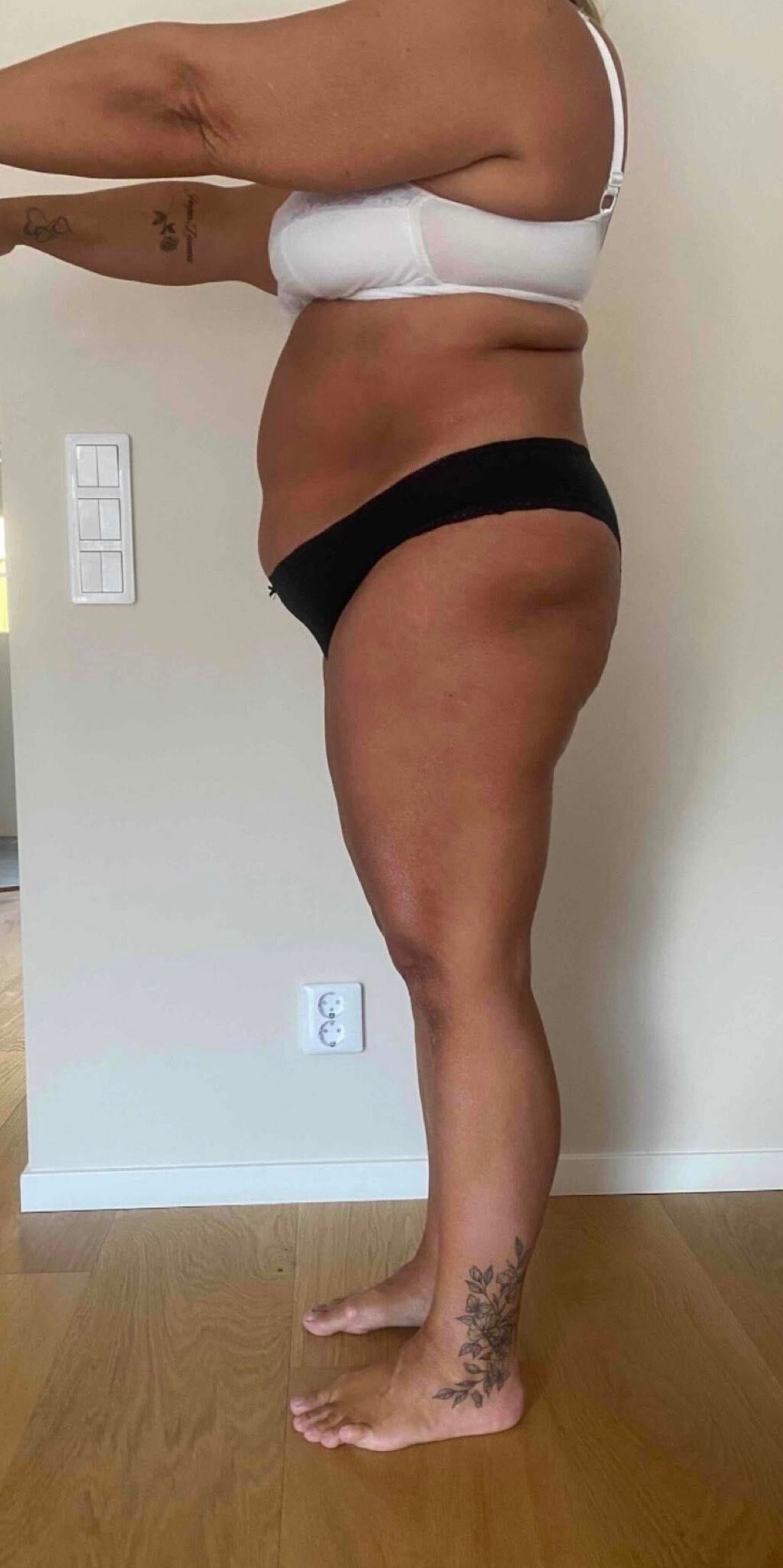 A person stands in profile against a plain wall, wearing a white bra and black underwear for a fitness progress check.
