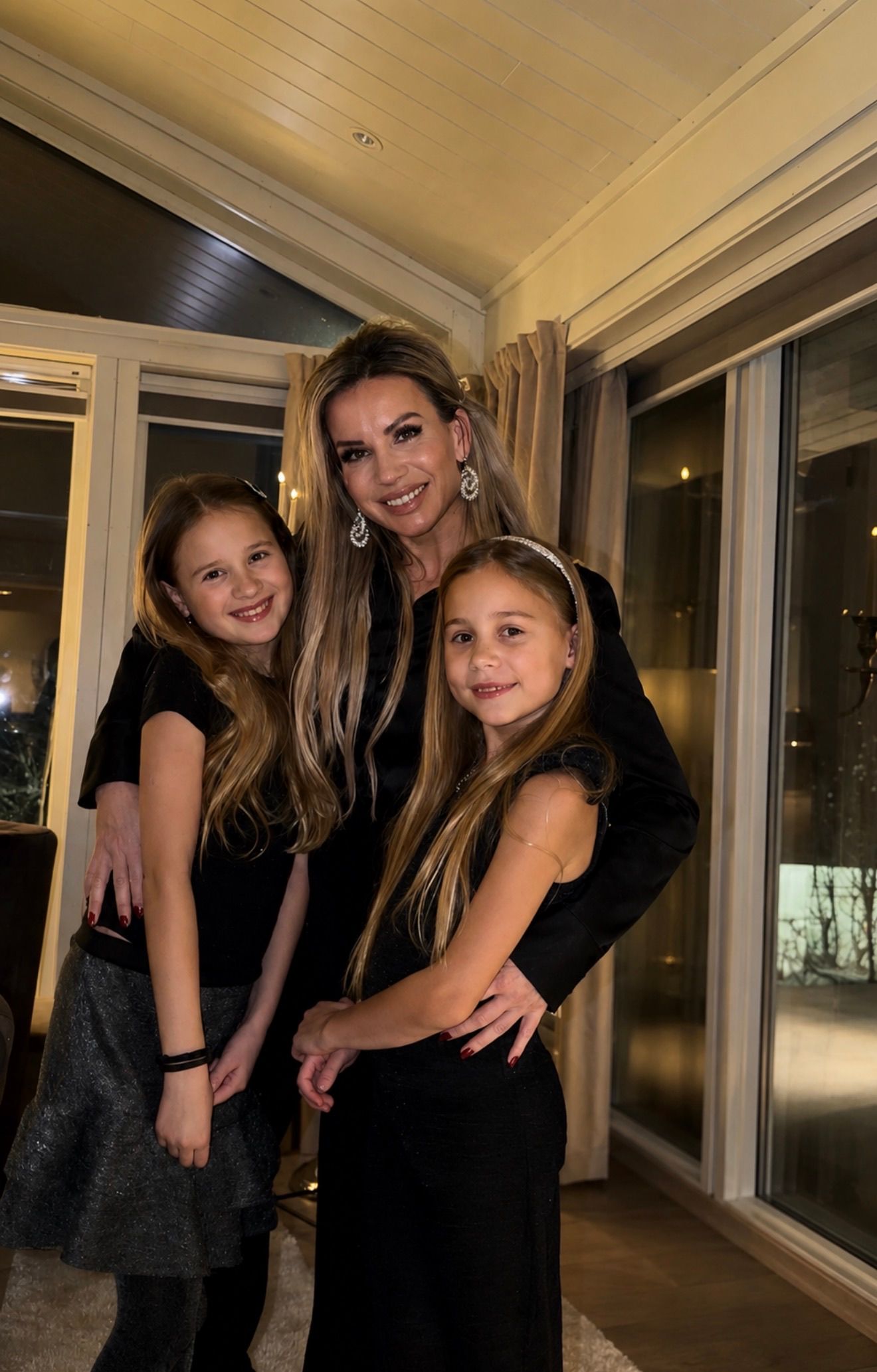 A woman smiling while embracing her two young daughters indoors.