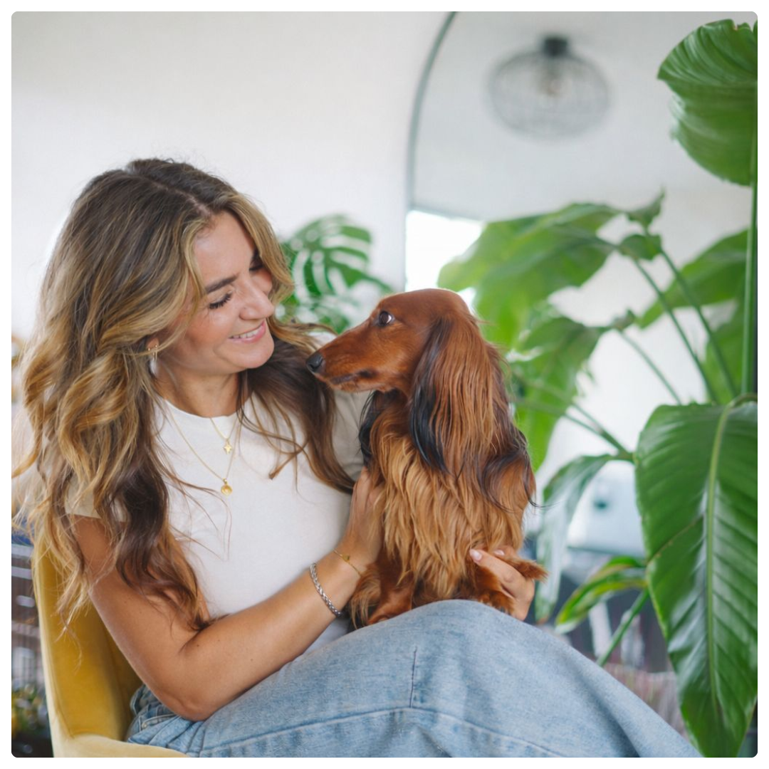 A woman with long wavy hair smiles while holding a long-haired dachshund in a bright room with large green plants.