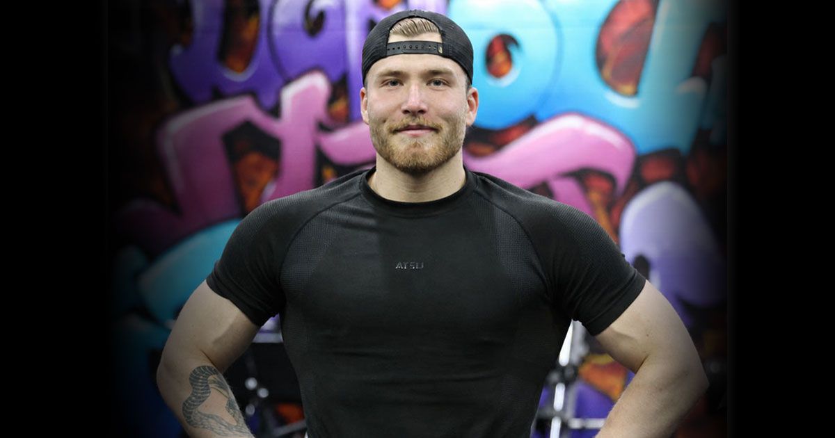 A muscular man with a beard and a black cap stands confidently in front of a colorful graffiti wall.