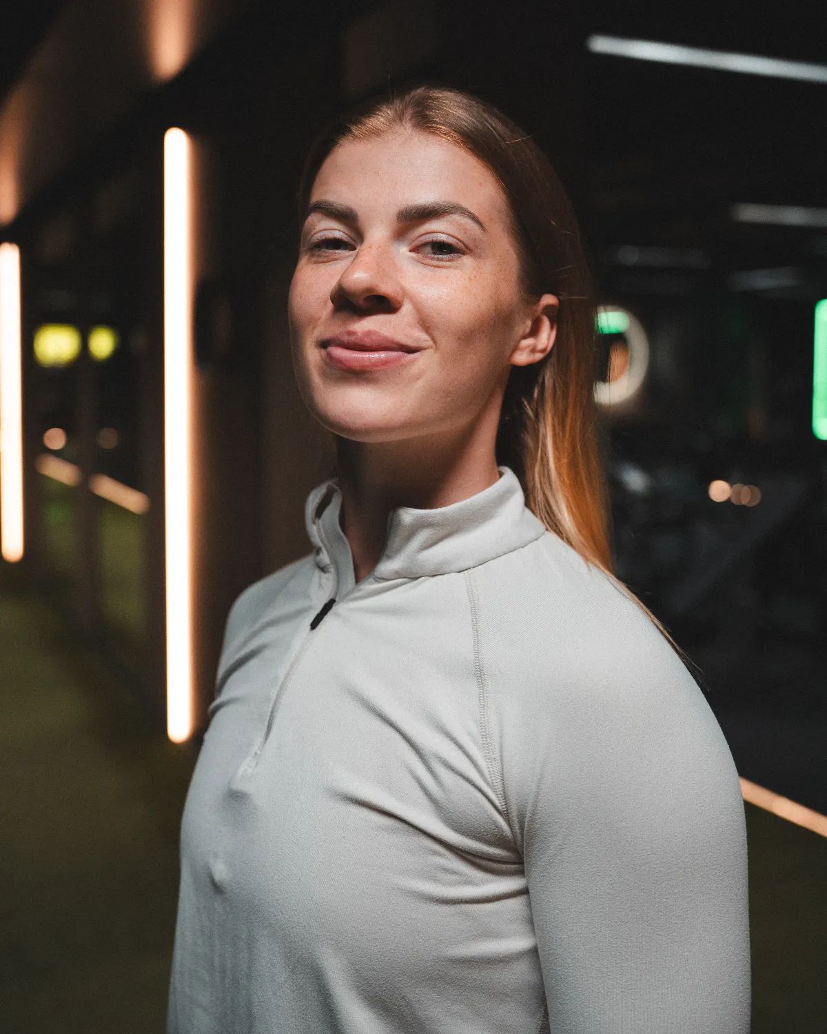 A woman with long blonde hair wearing a light-colored athletic top stands in a dimly lit gym.