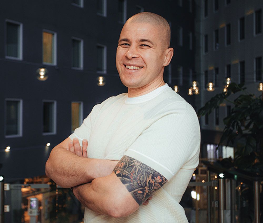 A smiling, bald man with a tattoo on his arm stands with his arms crossed in a modern building lobby.