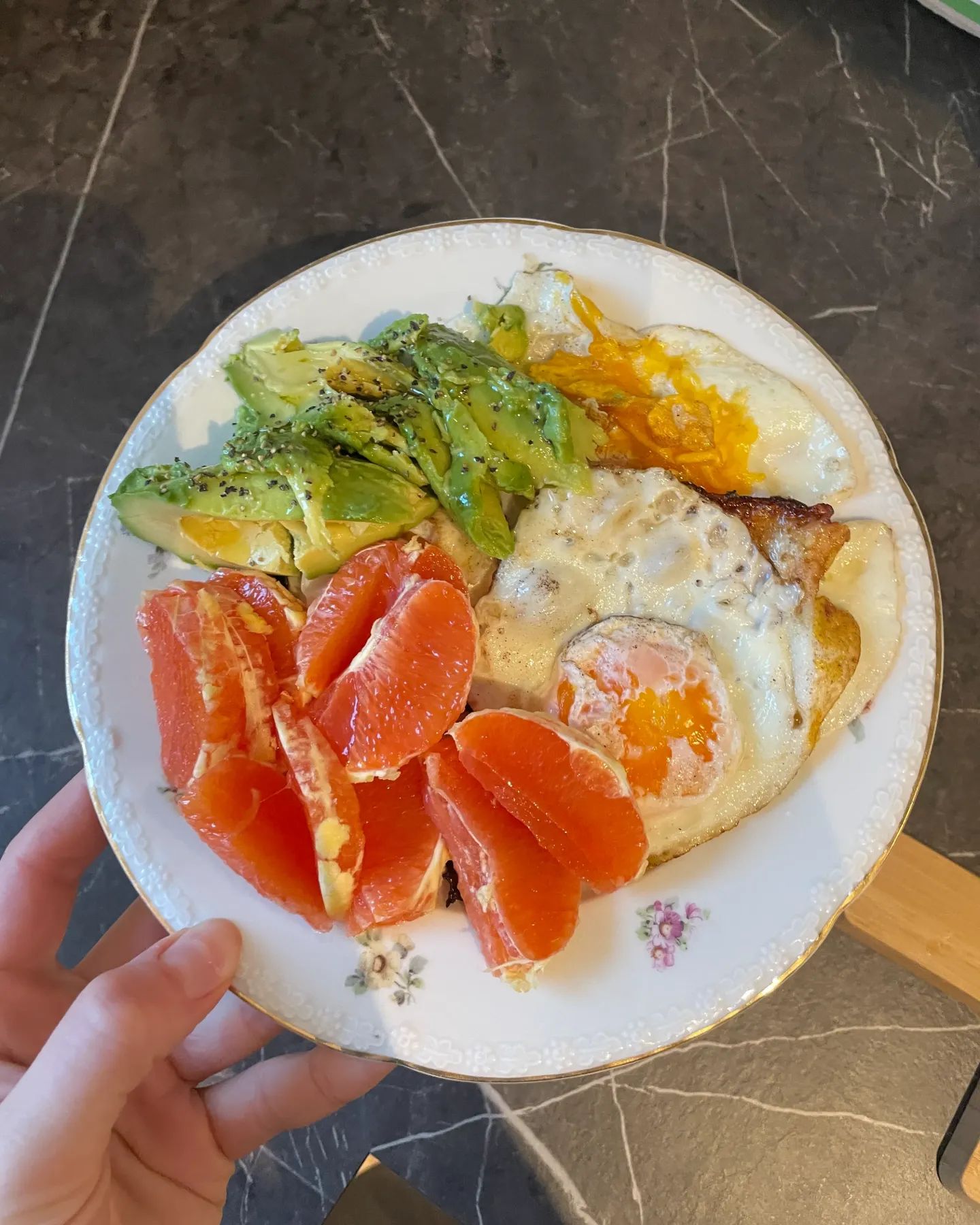 En hand håller en tallrik med avokado, stekt ägg och grapefrukt.