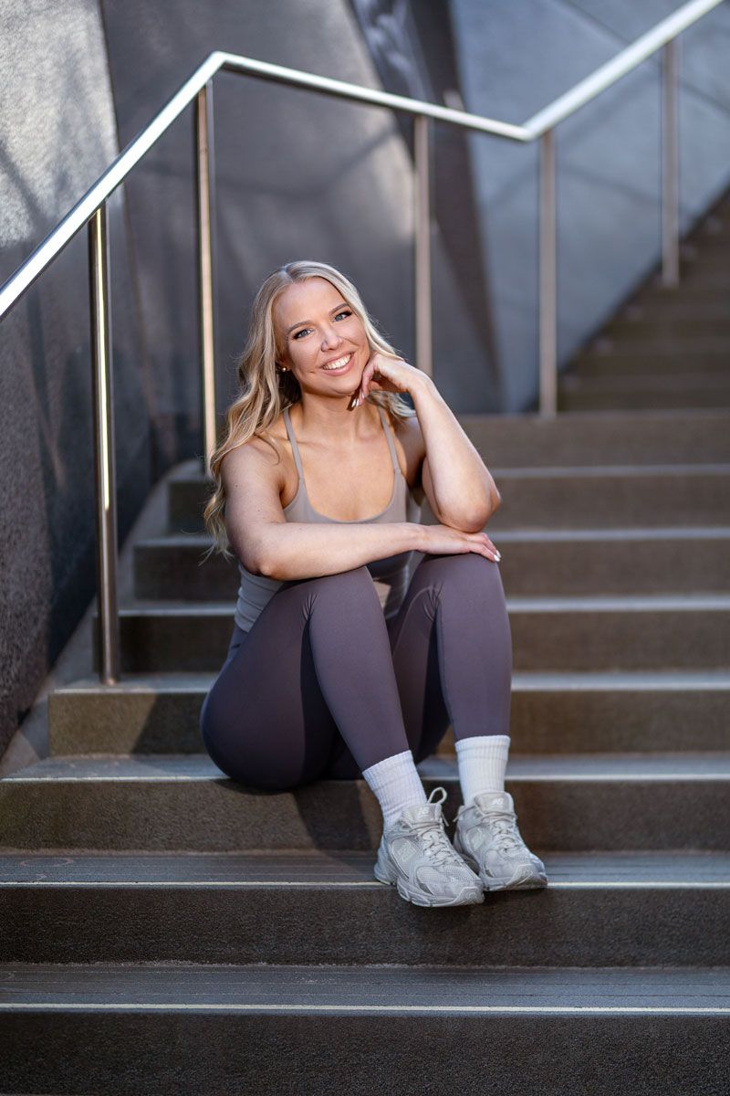 A smiling woman in athletic wear sits on outdoor concrete stairs with her hand resting on her chin.