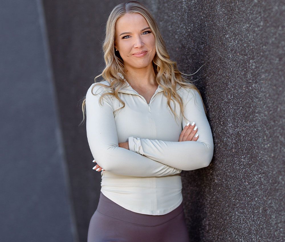 A blonde woman in athletic wear stands with her arms crossed, leaning against a dark textured wall.