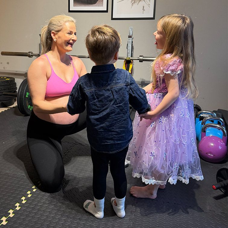 Gravid kvinne i treningsklær smiler til to barn i et treningsrom med vekter og kettlebells.