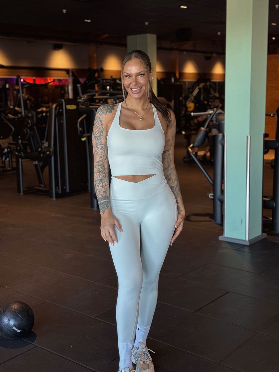 A smiling woman with tattooed arms stands in a gym wearing a matching light-colored athletic top and leggings.