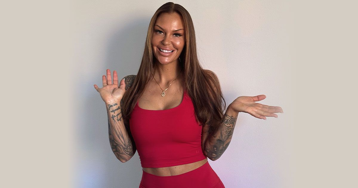 A smiling woman with long brown hair and arm tattoos wearing a red workout set poses with her hands raised.
