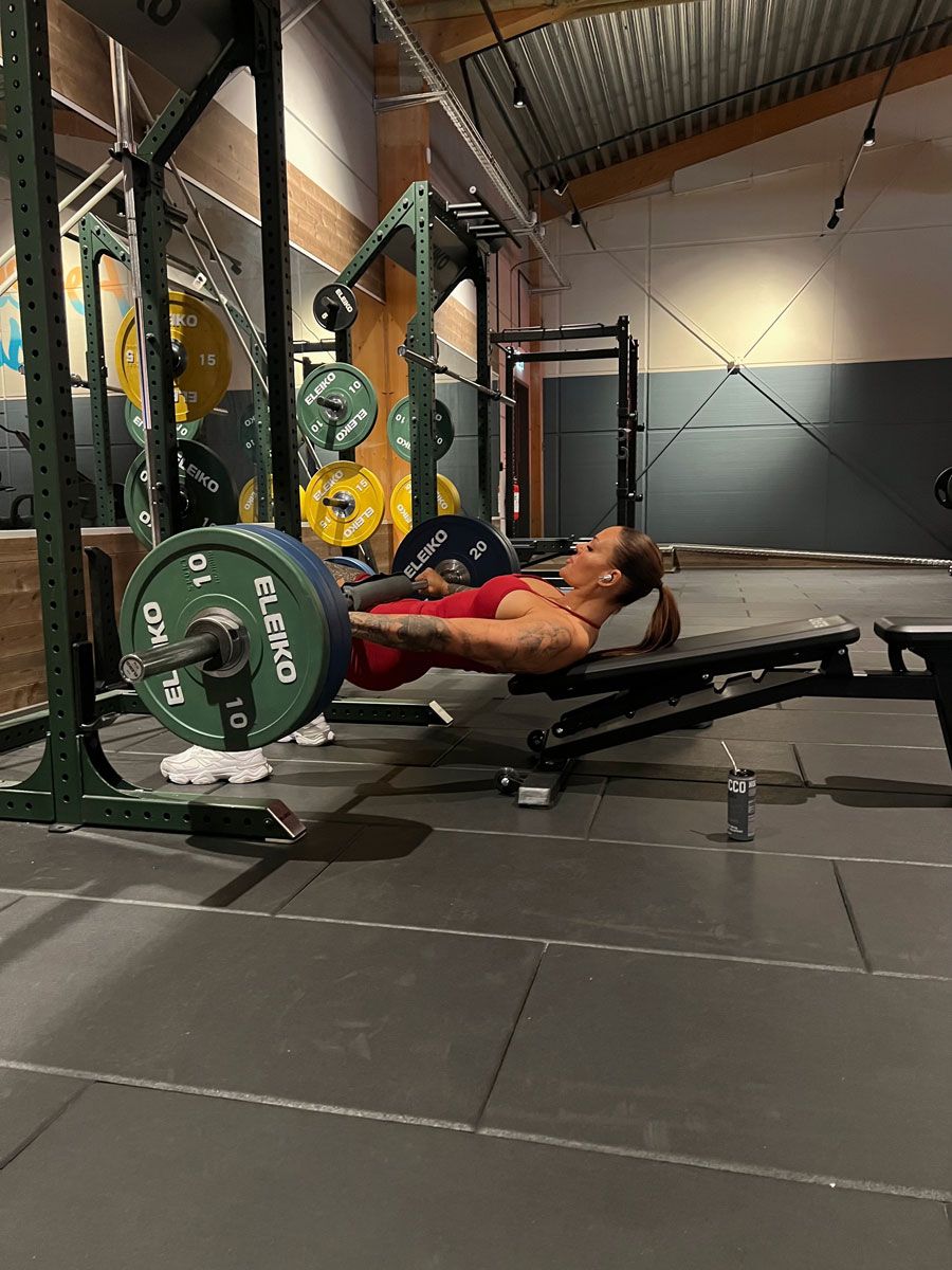 A woman in a red workout outfit performs a barbell hip thrust on a weight bench in a gym.