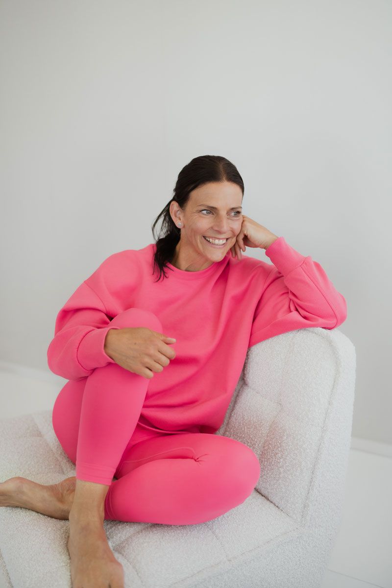 A smiling woman in a bright pink matching set sits comfortably on a white textured chair against a plain wall.