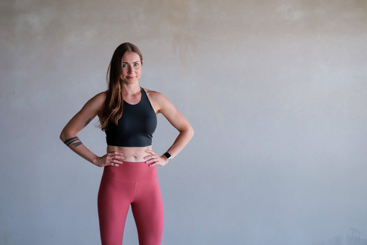 Maren Naue in schwarzem Sport-Top und roséfarbenen Leggings, Hände in den Hüften, vor einer grauen Wand