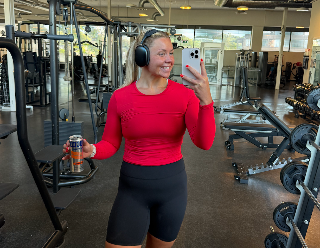 A smiling woman in a red long-sleeve shirt and black shorts takes a mirror selfie while holding a drink in a gym.