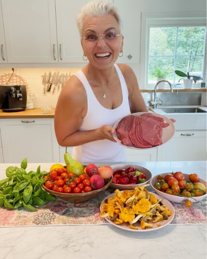 Camilla i sitt kök med en tallrik kött, framför skålar med tomater, frukt, paprika, basilika och kantareller
